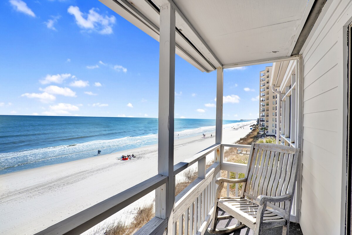 An oceanfront balcony is presented with a wooden rocking chair and an unobstructed view of the sandy beach and sparkling ocean waves. Bright blue skies and scattered clouds enhance the serene coastal setting.