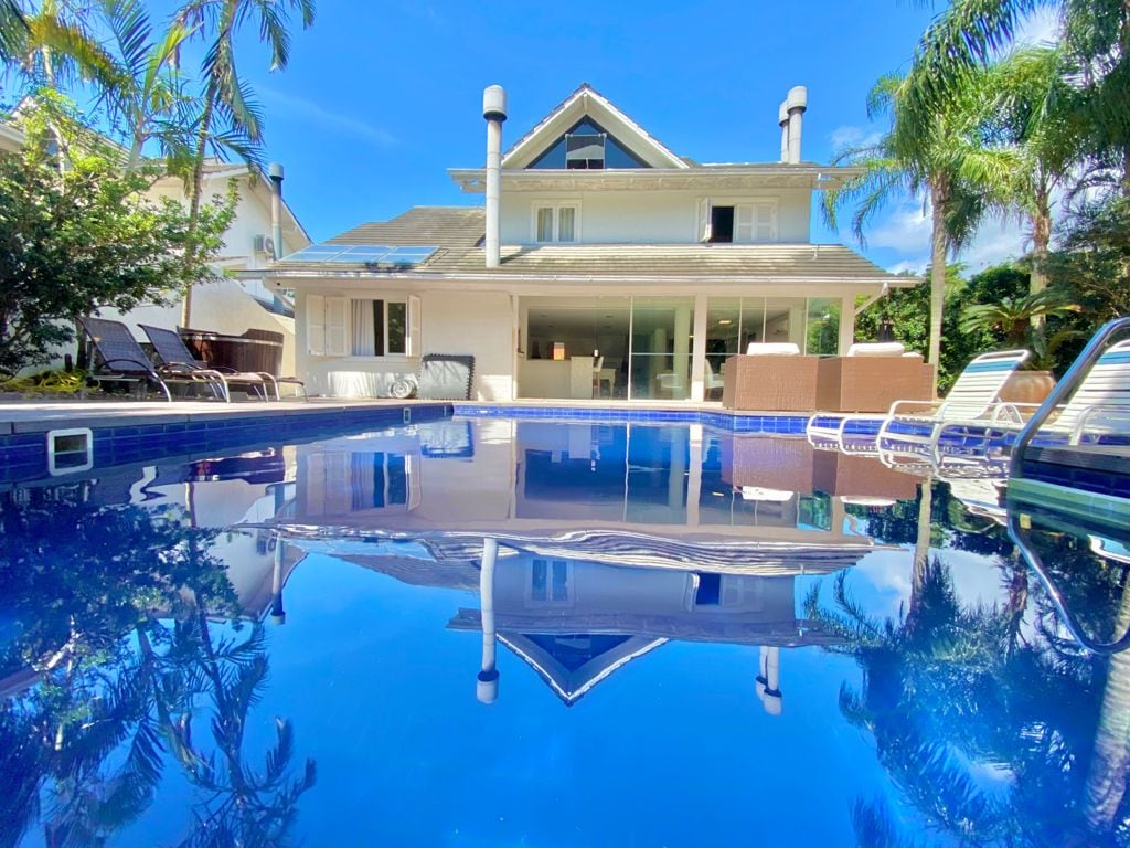 A serene view of a swimming pool reflects the two-story house with large glass doors. Surrounding the pool, sun loungers provide a space for relaxation, framed by lush greenery and a clear blue sky.