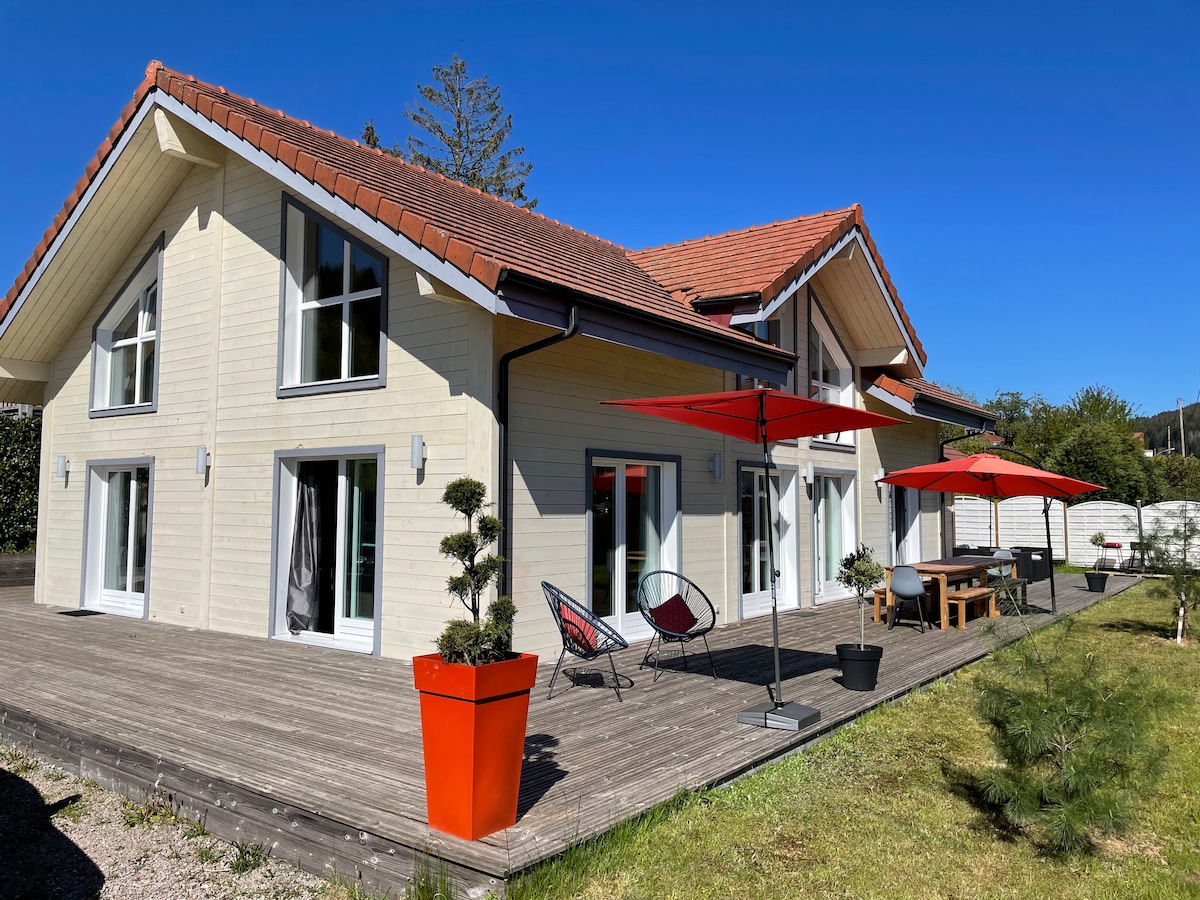 The modern chalet features a spacious wooden terrace, accented by bright red umbrellas and potted plants. Large glass doors allow natural light to fill the interior, while the symmetrical roofline adds architectural interest against the clear blue sky.