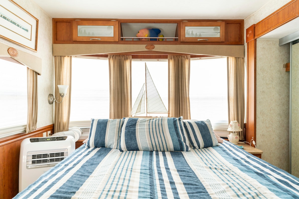 A spacious sleeping area is highlighted by a striped bedspread, complemented by light streaming in from large windows with sheer drapes. Above the bed, a decorative shelf displays a small figurine, and a nightstand is positioned beside the bed.