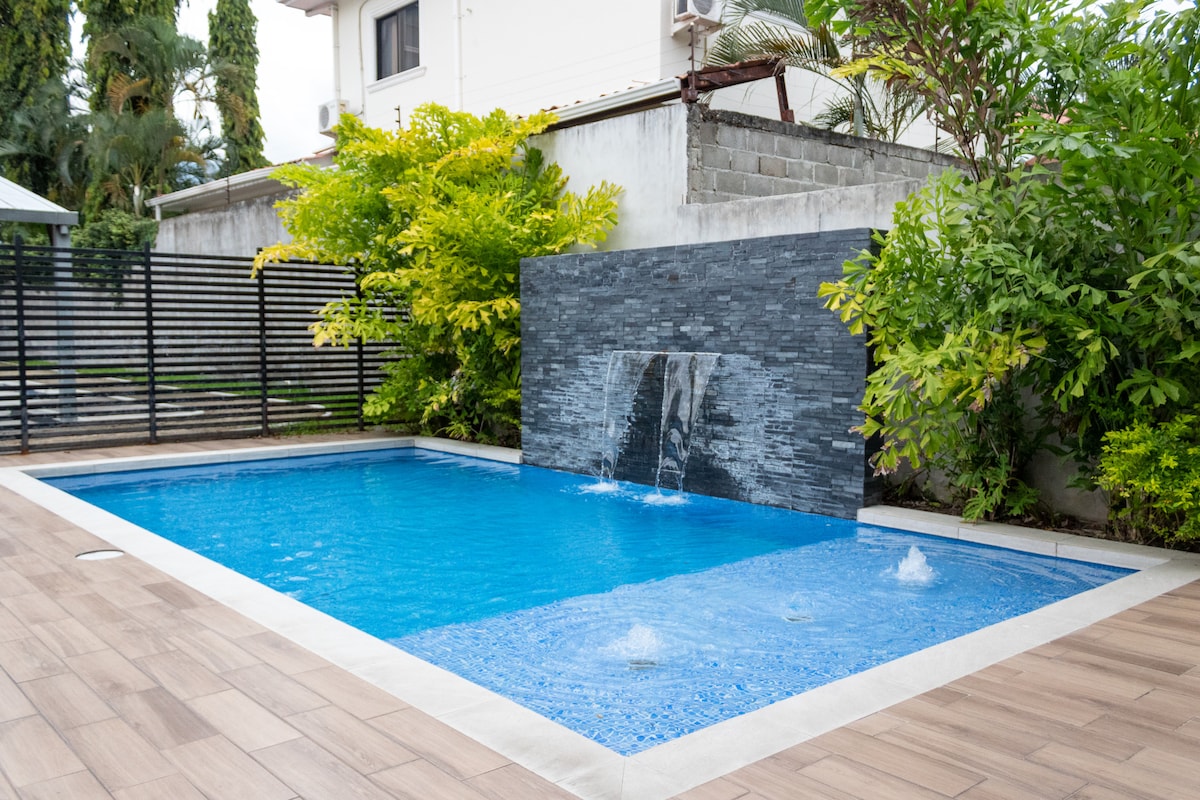 A swimming pool is surrounded by a wooden deck and features a stone water wall that adds a stylish touch. Lush greenery frames the space, creating a tranquil environment. Water fountains are integrated into the pool, providing a soothing ambiance.