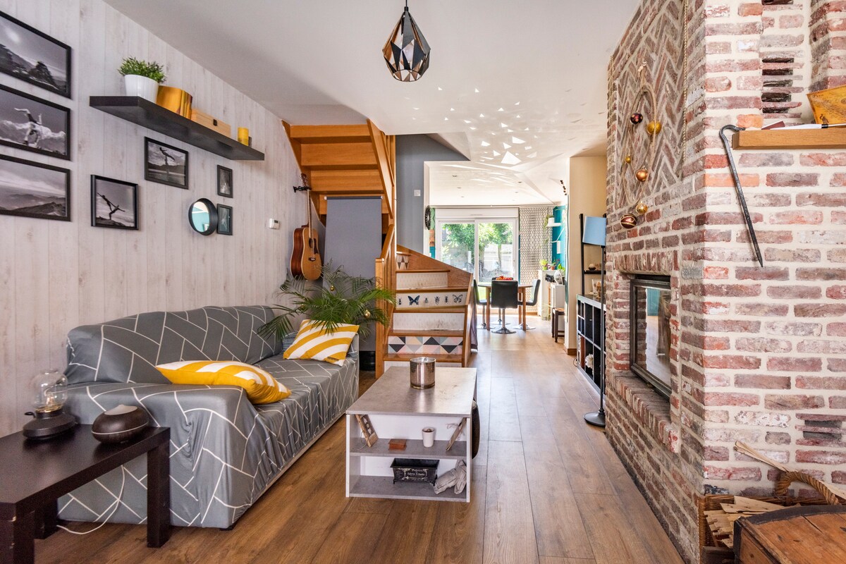 A stylish living area features a light gray sofa adorned with vibrant yellow cushions. A wooden coffee table sits in front, accompanied by artfully arranged photographs on the walls. Sunlight filters through large windows, illuminating the open space that leads toward a dining area.