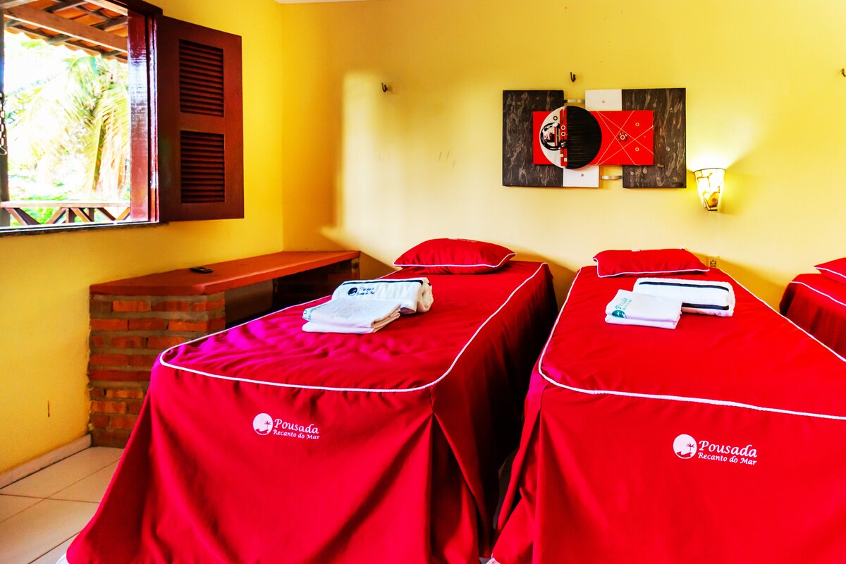 The room features two twin beds dressed in red linens, each topped with neatly arranged towels. A natural light flows through the wooden window, enhancing the soft yellow walls. A wooden bench is positioned against the wall, contributing to the room's functional layout.
