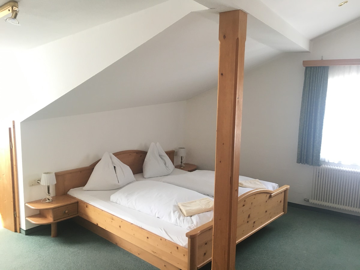 A spacious sleeping area is characterized by a wooden bed featuring two soft pillows and a light blanket. Soft lighting is provided by bedside lamps, and a window allows natural light to brighten the room. Traditional wooden furniture complements the serene environment.