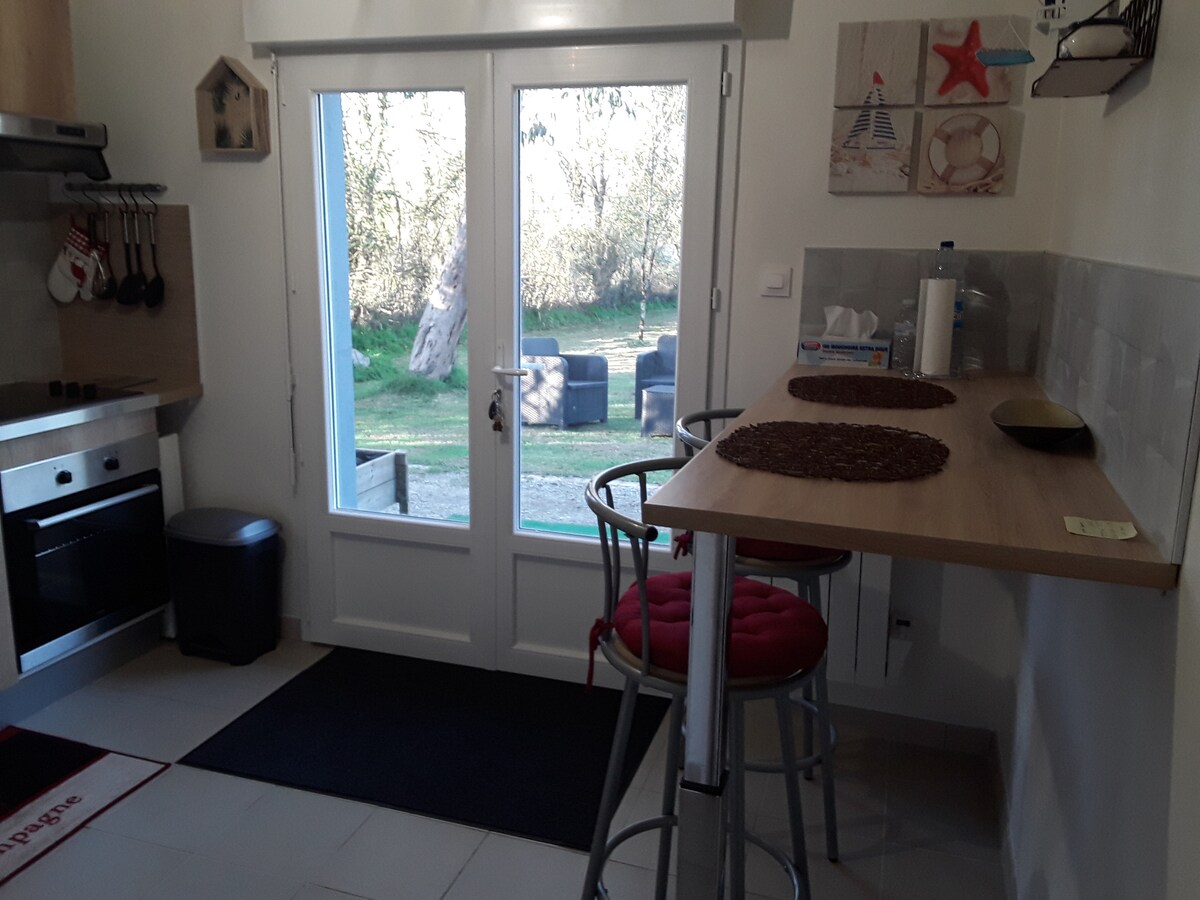 A kitchen area features a countertop with two barstools, overlooking a garden view through large glass doors. Kitchen appliances include an oven and a coffee machine. Natural light brightens the space, while decorative elements add a touch of warmth.