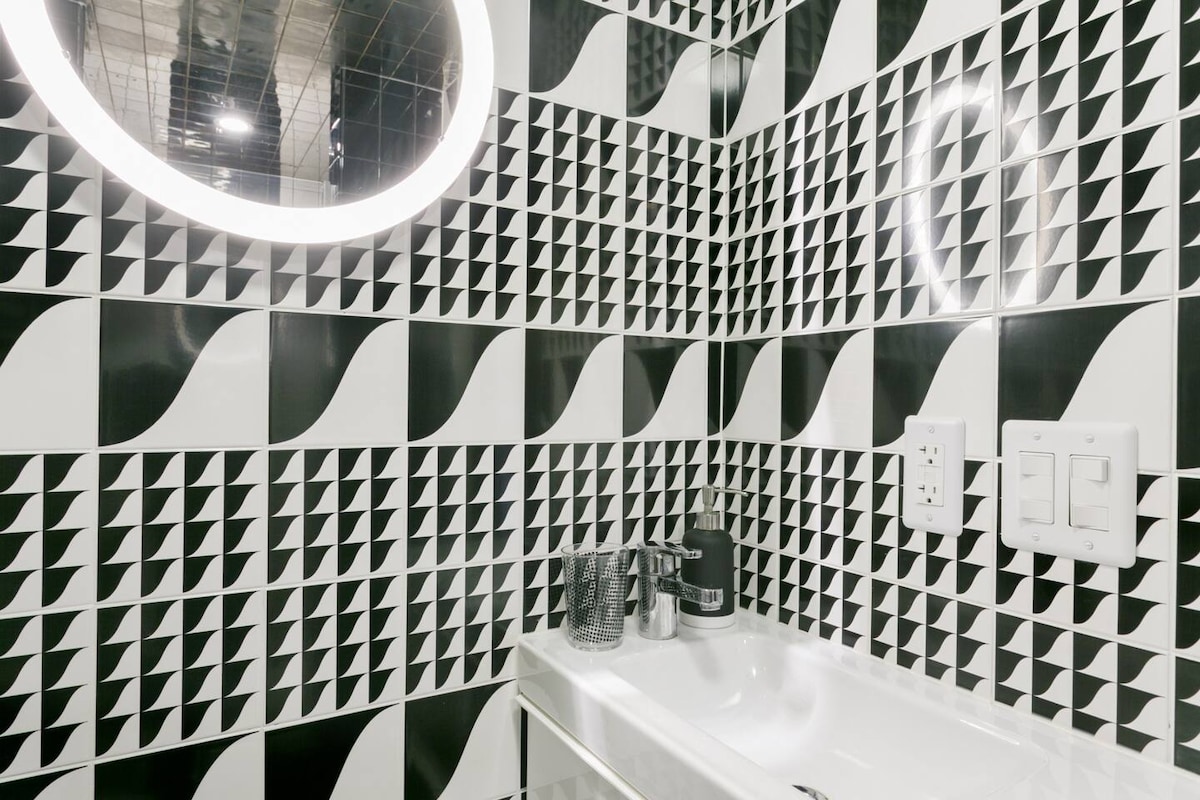 A modern bathroom features eye-catching black and white geometric tiles that create a bold visual impact. A round illuminated mirror enhances the sleek design above a clean, white sink, with minimalist toiletries arranged neatly nearby.
