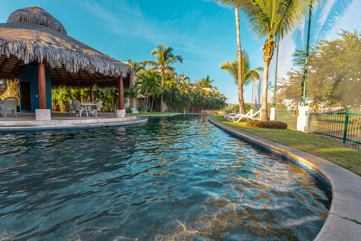 A sprawling swimming pool is set within a lush garden, featuring a traditional palapa with seating. Tall palm trees surround the area, creating a serene environment. Clear blue water reflects the sky, inviting guests to relax and enjoy the tranquil setting.