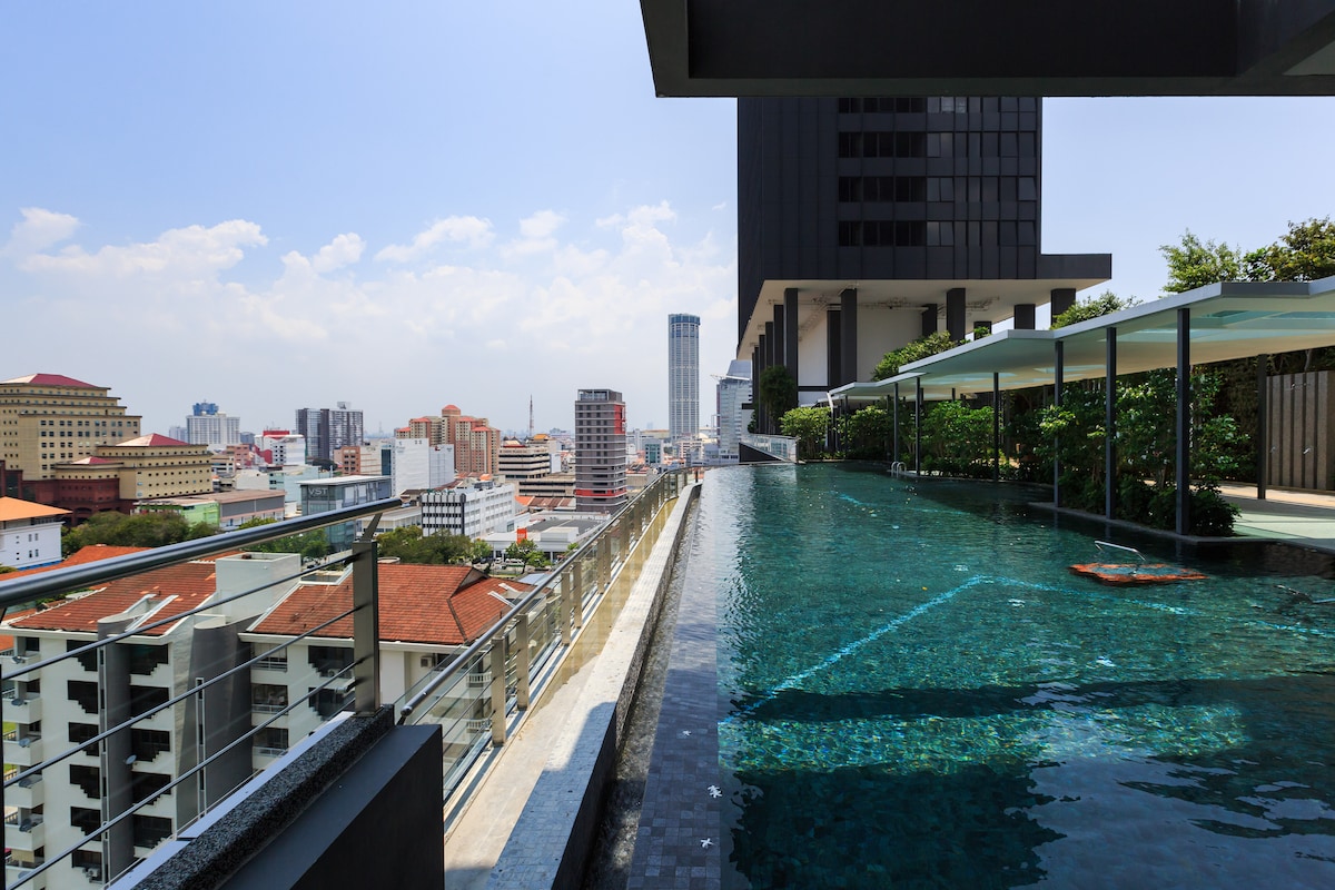 An infinity-edge swimming pool is viewed from the edge of a rooftop, presenting a skyline with a mix of residential and commercial buildings in the background. The pool area is surrounded by greenery, with lounge chairs and shaded spaces available for relaxation.