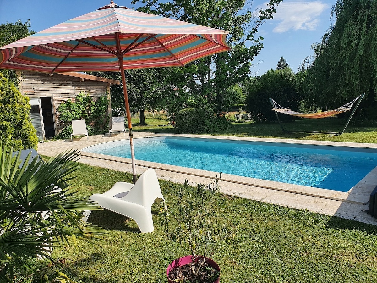 A refreshing outdoor pool is set against a backdrop of lush greenery. A colorful striped umbrella offers shade over a white lounging chair, while a hammock can be seen nearby. A potted plant adds a touch of vibrancy to the inviting pool area.