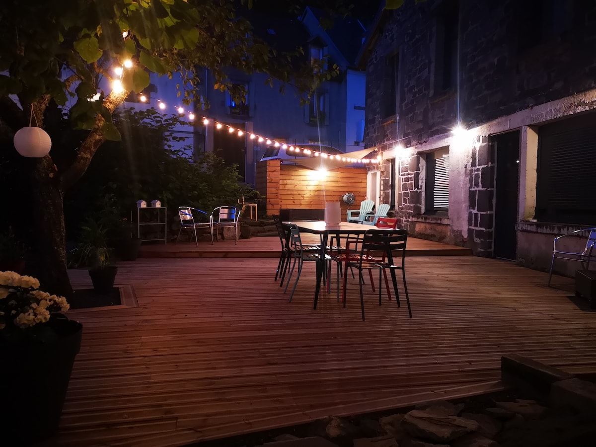 An outdoor terrace is illuminated by decorative string lights, creating a warm ambiance. A wooden deck features a dining table surrounded by colorful chairs, with additional seating visible nearby. Lush greenery offers a natural backdrop to the cozy gathering space.
