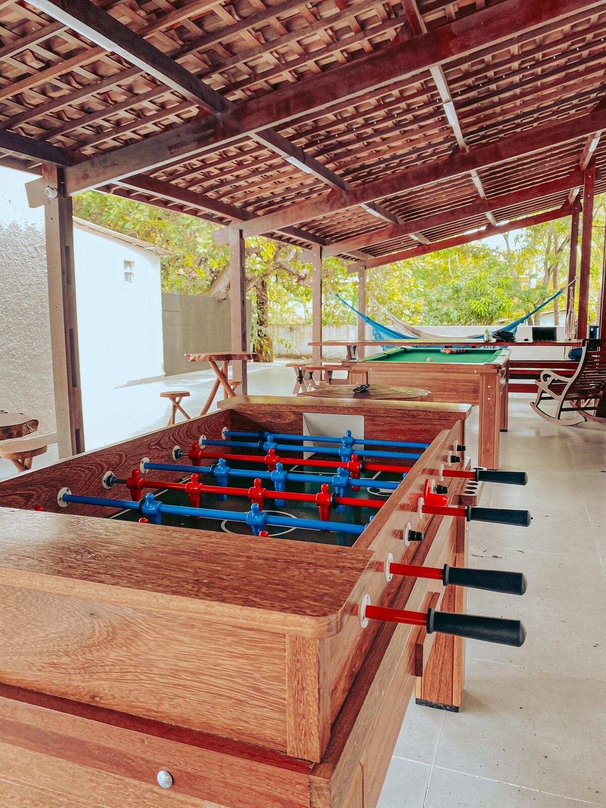 An outdoor recreational area is showcased, featuring several foosball tables made of dark wood. The space is shaded by a traditional roof, with natural light filtering in. Nearby, a hammock is visible, complemented by wooden seating and tables under lush greenery.