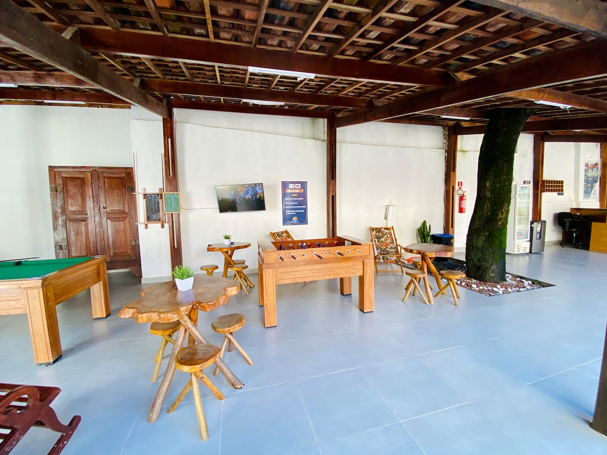 A communal area is presented, featuring a pool table and a foosball table. Wooden tables and chairs are arranged for seating. Natural light illuminates the space, complemented by a large tree positioned centrally, adding a touch of greenery to the room.
