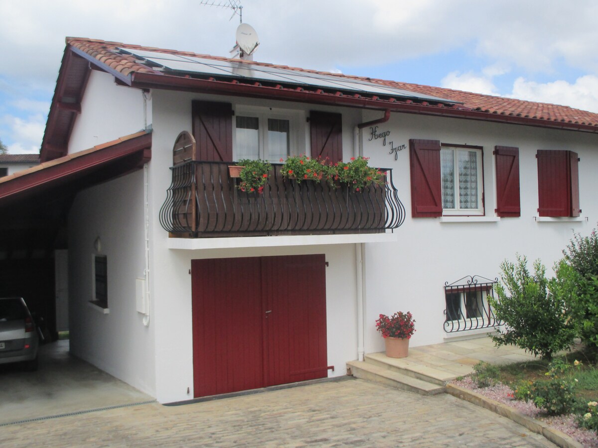 A two-story house with a white facade and red shutters features a covered parking area below. The balcony adorned with flower boxes enhances the exterior, while a well-maintained garden with shrubs adds greenery to the front of the property.