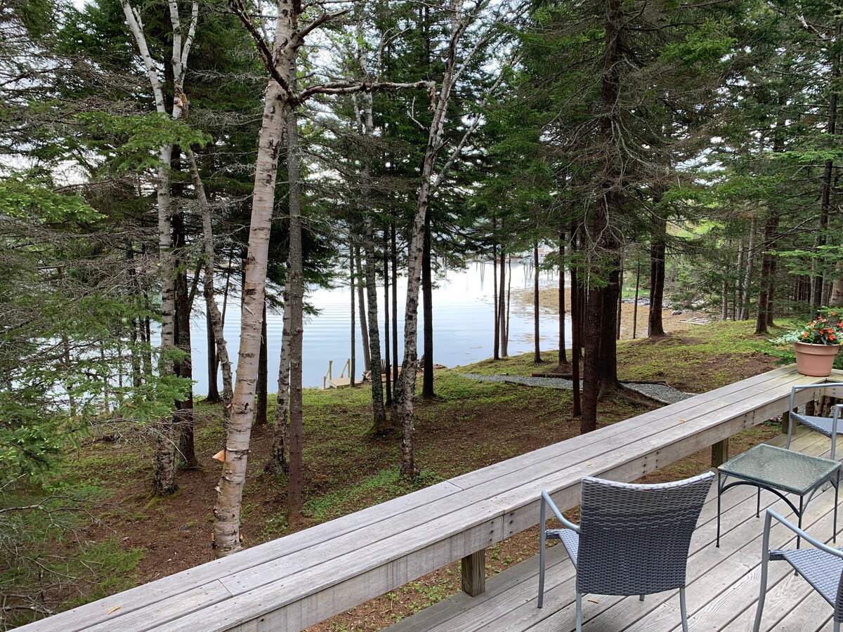 A wooden deck extends along the side of the cottage, looking out towards calm waters framed by tall trees. Two metal chairs with cushions are positioned on the deck, inviting visitors to enjoy the serene view of the harbor area and surrounding nature.