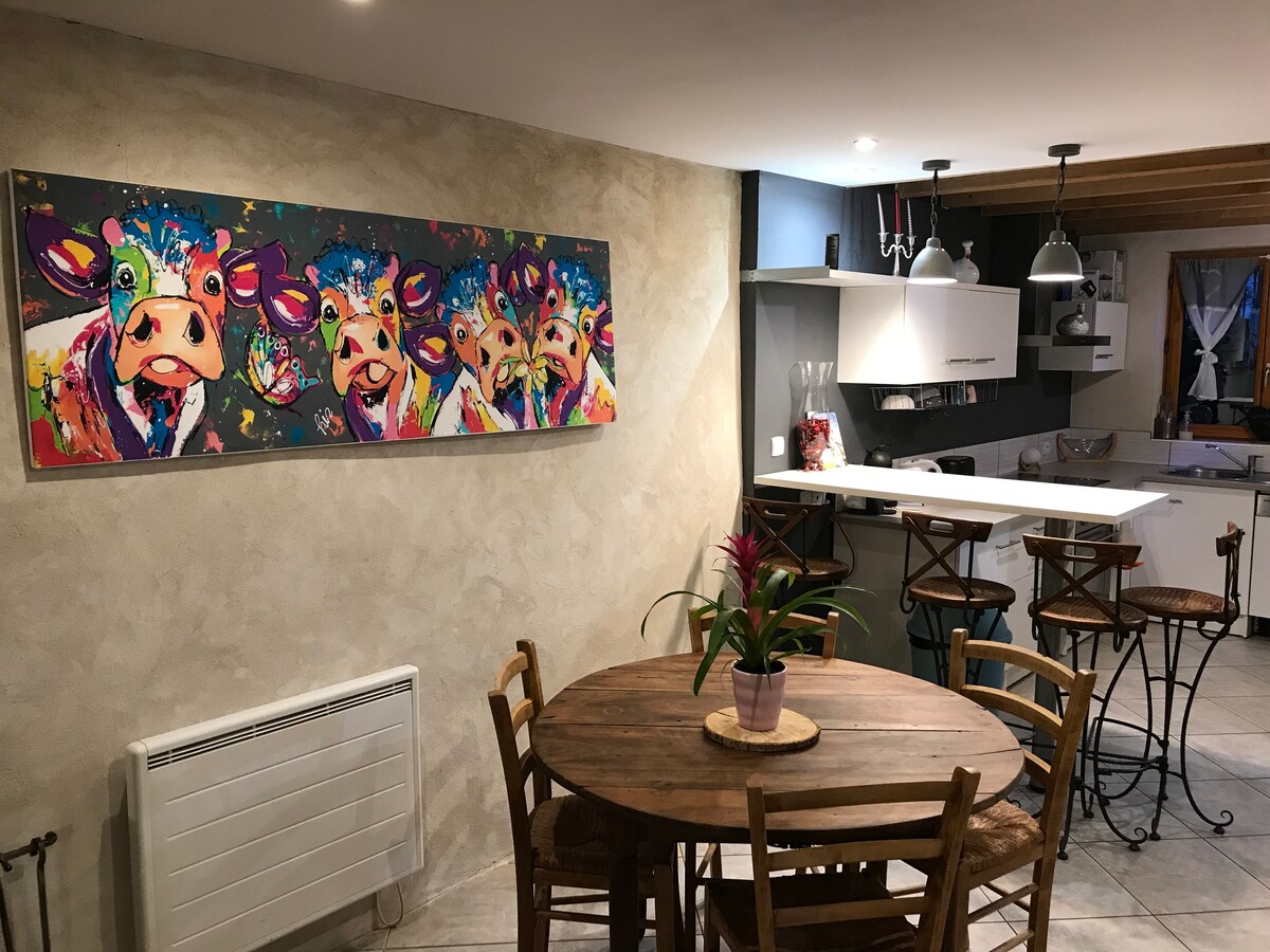 A dining area features a round wooden table surrounded by wooden chairs. A colorful wall art piece depicting cows adds a playful touch. The kitchen provides a modern, open layout with bar seating, illuminated by pendant lights.