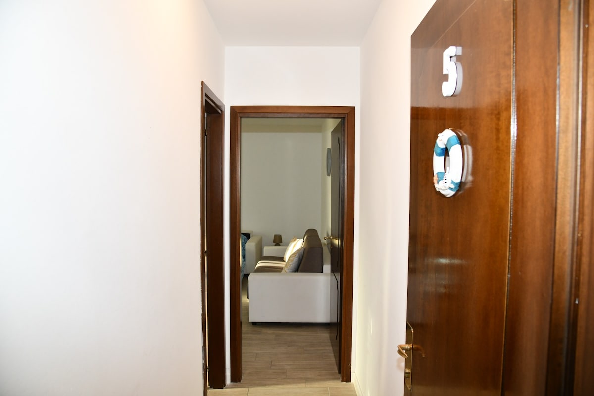A well-lit hallway leads towards an open doorway, providing a glimpse into a living area. The floor is tiled in light colors, and white walls enhance the spacious feel. The door on the right features a wooden finish with a decorative number, indicating the unit number.