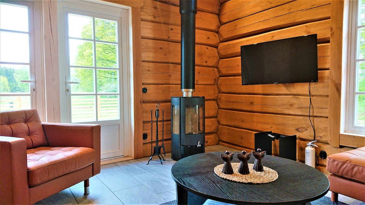 A cozy living area features a wood-burning stove against a backdrop of log walls. A flat-screen television is mounted on the wall opposite a round coffee table adorned with decorative items. Two brown leather armchairs provide additional seating, creating a welcoming space.