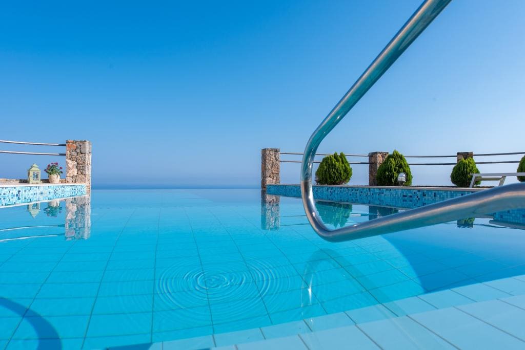 A clear blue swimming pool is framed by stone edges, with stainless steel handrails leading into the water. Lush greenery is visible on the surrounding walls, contrasting with the vibrant aqua tiles of the pool, against a backdrop of bright blue sky.