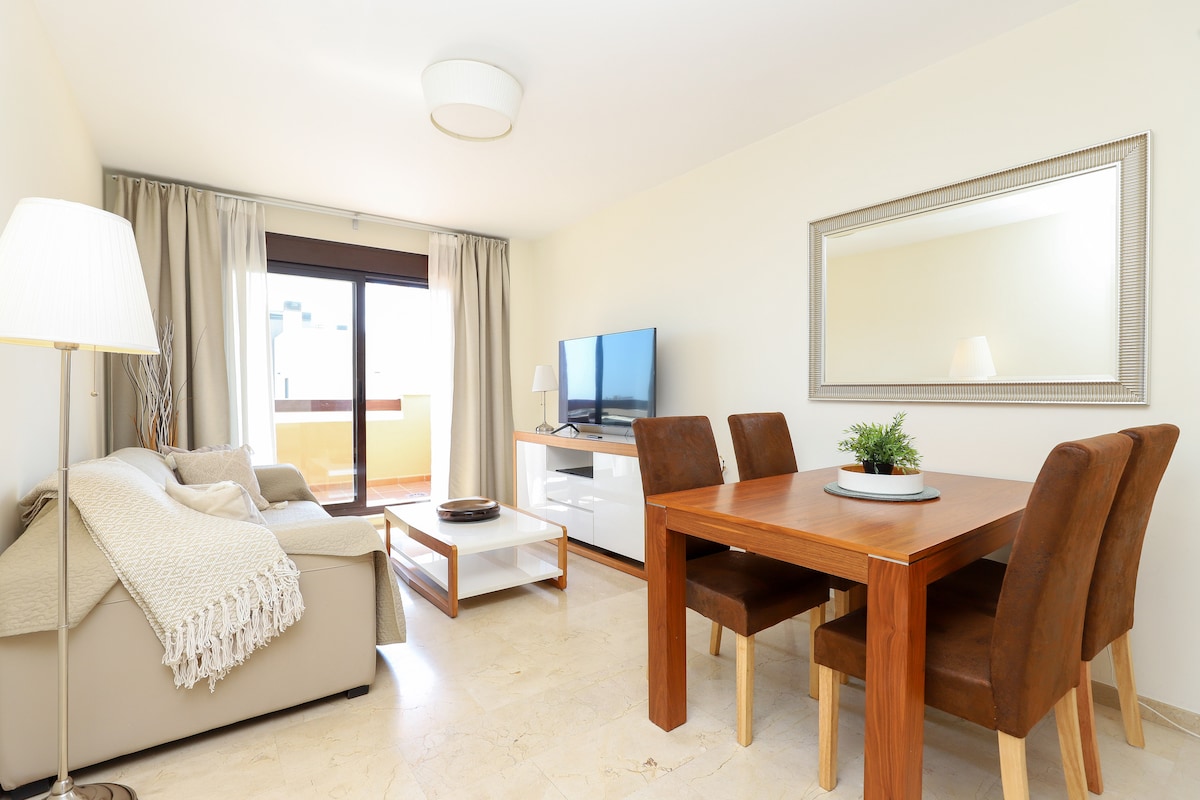 A comfortable living space is featured, showcasing a light-colored sofa draped with a knitted throw. A wooden dining table with four chairs sits adjacent, complemented by a flat-screen TV mounted on a white console. Natural light filters through the sliding glass doors leading to a balcony.