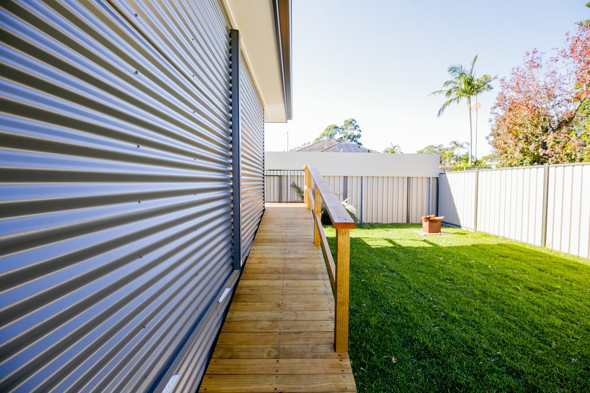 A wooden ramp extends from the entrance, leading into a grassy backyard area. The exterior features a sleek, corrugated metal design, and greenery from surrounding trees adds a natural touch. The space offers a pathway to enjoy the outdoors with ease.