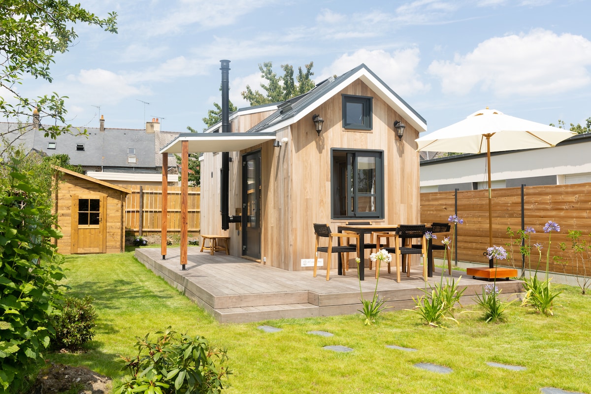 The tiny house is surrounded by a manicured garden, featuring a wooden deck and a small seating area with a table and chairs. A sun umbrella offers shade, while a wooden shed and landscaped greenery complement the natural setting.