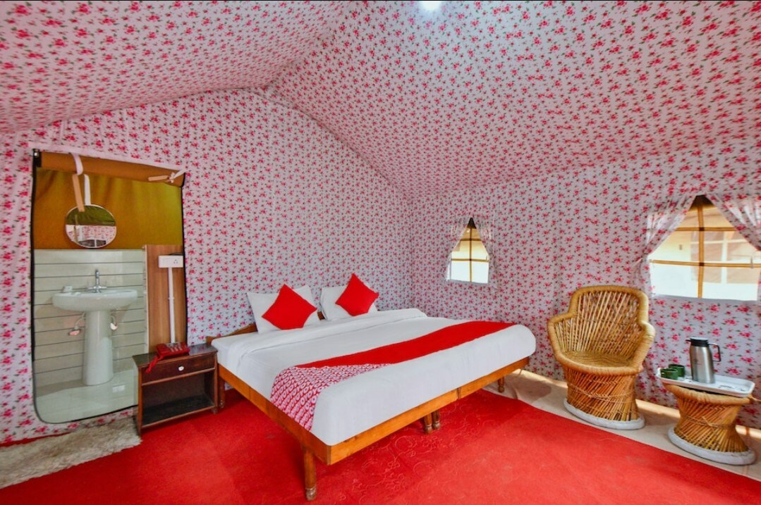 A cozy sleeping area is filled with floral-patterned walls and soft red accents on the bedding. A spacious bed is positioned at the center, with woven chairs placed nearby. Natural light filters in through small windows adorned with matching curtains.
