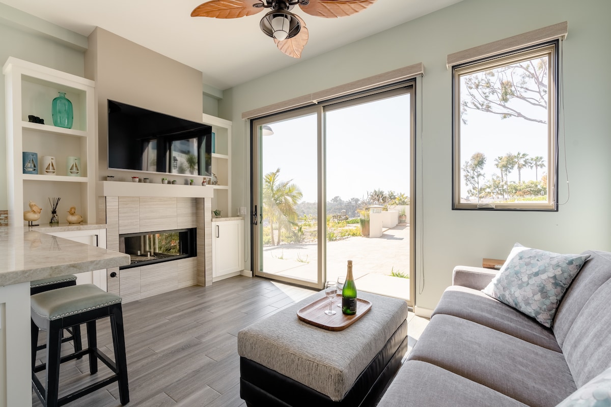 A spacious living area features large glass sliding doors leading to a patio, allowing natural light to fill the room. A comfortable sectional sofa is paired with a soft ottoman, and a wall-mounted television is positioned above a modern fireplace. Decorative shelves showcase various items.