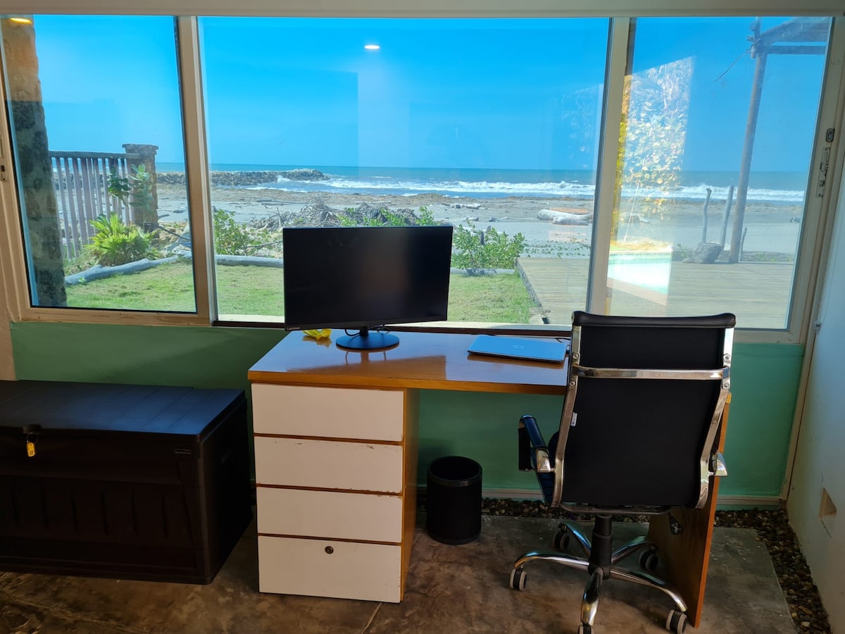 A workspace is featured with a sleek desk and ergonomic chair, offering a view of the ocean through large windows. A computer monitor sits on the desk, adjacent to a notepad. The tranquil beach scenery is visible outside, enhancing the work environment.
