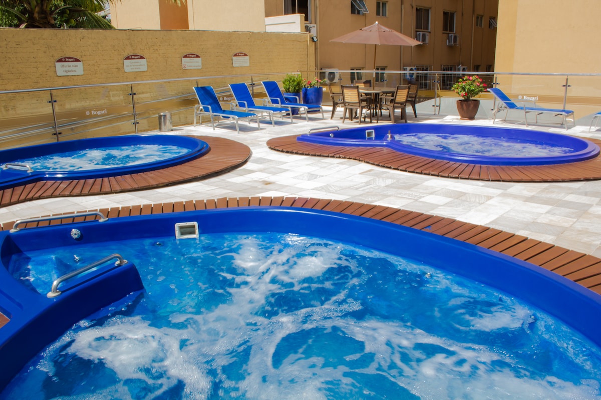 Three distinct hydro-spas are depicted, each featuring a vibrant blue color and surrounded by wooden decking. Comfortable lounge chairs are positioned nearby, accommodating relaxation. A dining table with chairs is situated under a large umbrella, enhancing the leisure atmosphere in the designated area.
