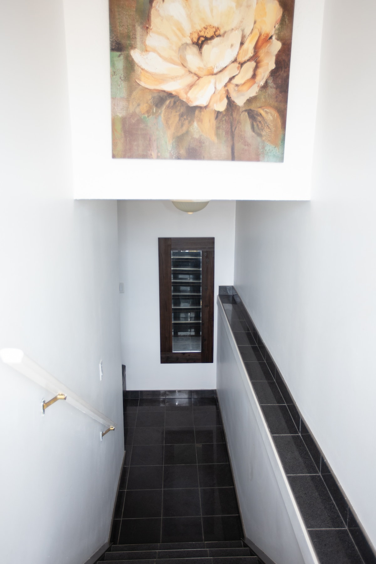 A staircase is visible leading downwards, flanked by white walls and a dark tile railing. Above, a floral art piece is displayed, while a multi-paneled mirror is positioned on the wall at the bottom of the stairs, enhancing the sense of space.