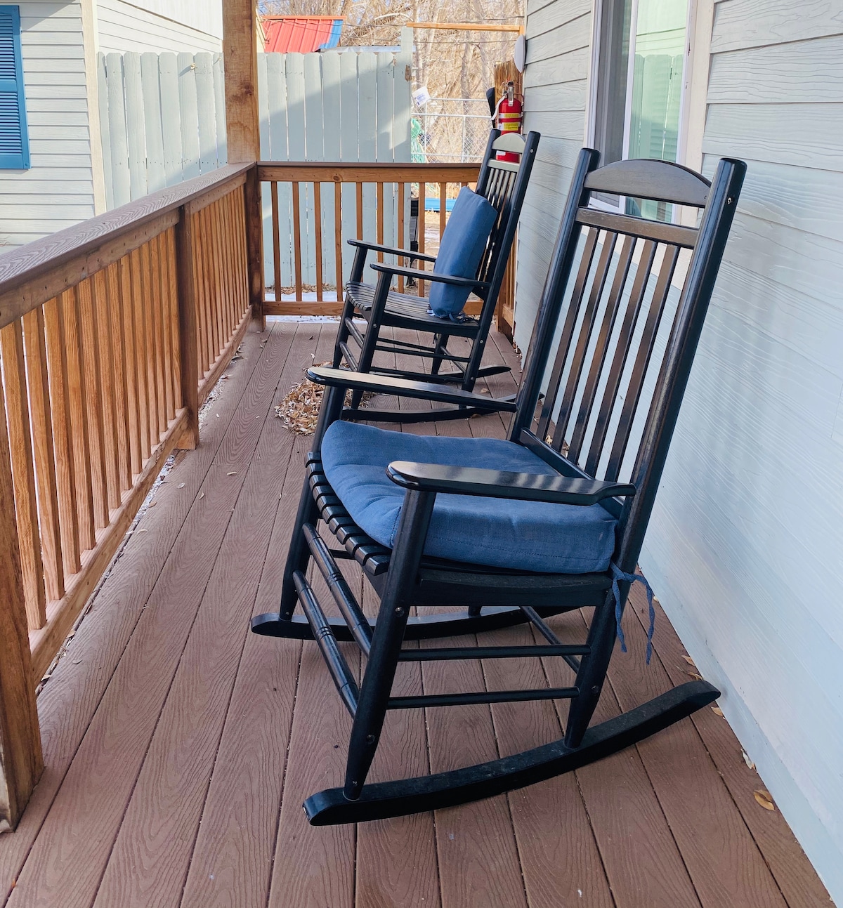 Two black rocking chairs with blue cushions are positioned on a wooden porch, offering a comfortable spot for relaxation. The porch railing features a warm wooden finish, and a portion of the exterior is visible behind the chairs.