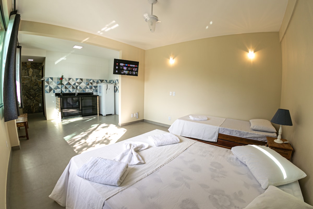A bright and spacious suite features two beds arranged comfortably across the room. A kitchenette area is visible in the background, with colorful tiles providing a lively backdrop. Large windows allow natural light to fill the space, complemented by simple, modern decor.