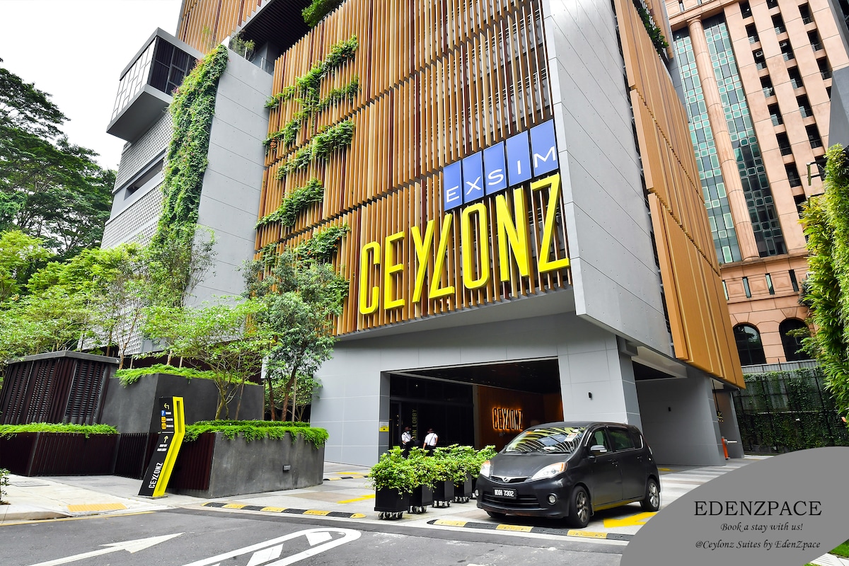A modern building exterior is shown with a clean design. Lush greenery is present on the facade, enhancing the appearance. An entrance is marked with a prominent sign displaying 'EXSIM CEYLONZ'. A parking area is visible, with a vehicle parked in front.