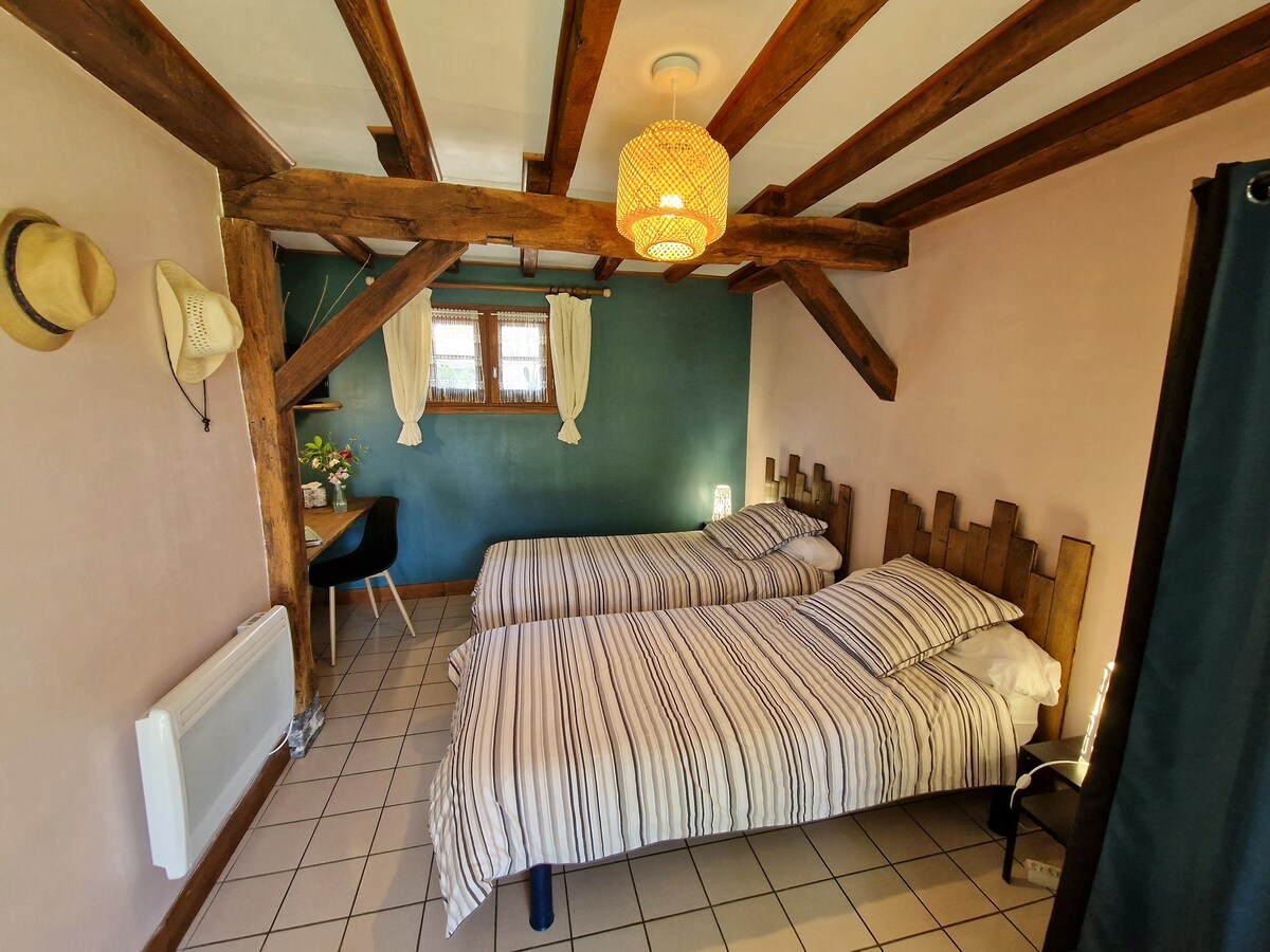 A cozy room features two single beds with striped linens, positioned alongside a wall adorned with wooden accents. A small desk and chair are situated under a window, while a pendant light hangs from the ceiling. Decorative elements include hats displayed on the wall.