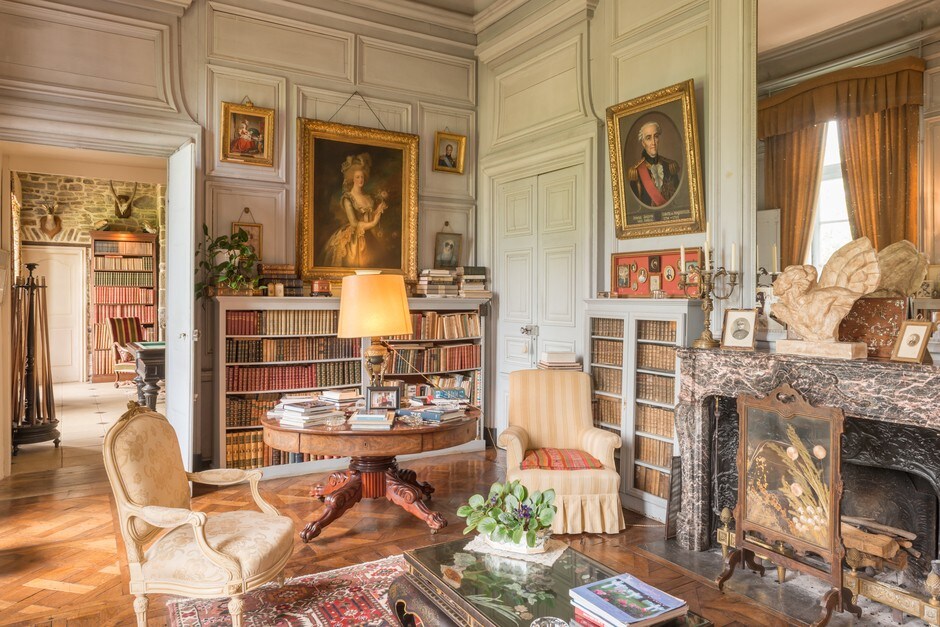 An elegant library features richly crafted wooden shelves filled with books, complemented by intricate decor and artwork. A classic round table is accentuated by a table lamp, and comfortable seating is arranged nearby. Light pours in from tall windows, highlighting the historic ambiance.