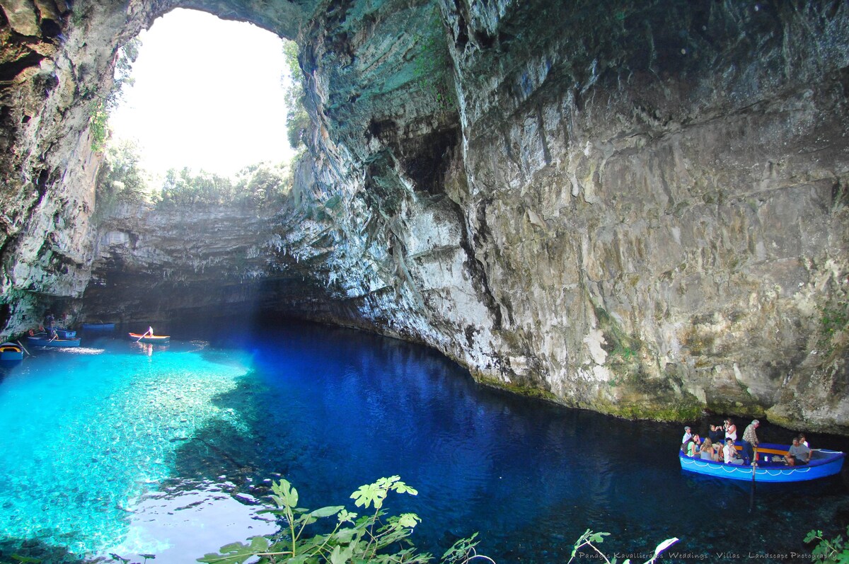 A natural cave with high rocky walls is illuminated by an opening above, revealing a vibrant blue lake below. Small boats float on the water, with visitors enjoying the serene surroundings. Lush green foliage is visible at the foreground, enhancing the cave's natural beauty.