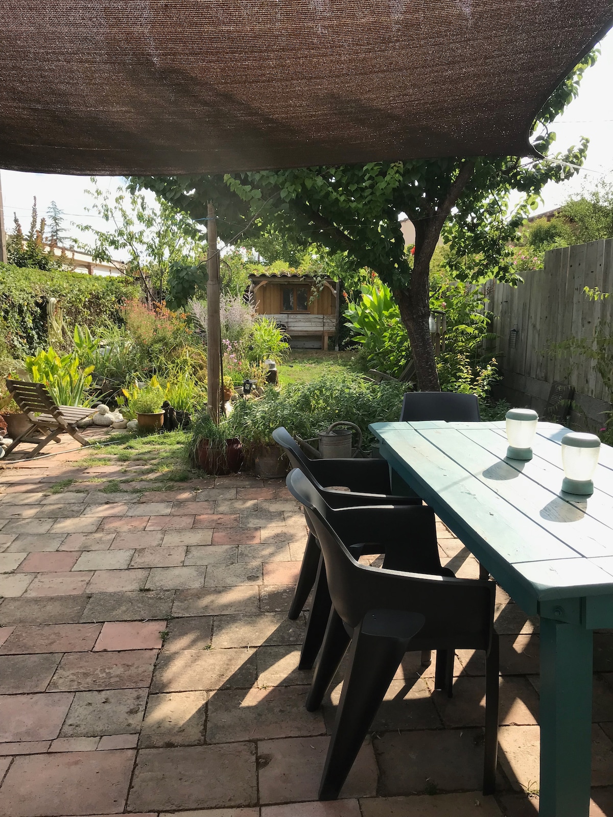 A sunlit terrace features a wooden table accompanied by four black chairs, surrounded by a lush garden. Various plants and greenery are visible, along with a shaded area provided by a large canopy. A wooden structure can be seen in the background.