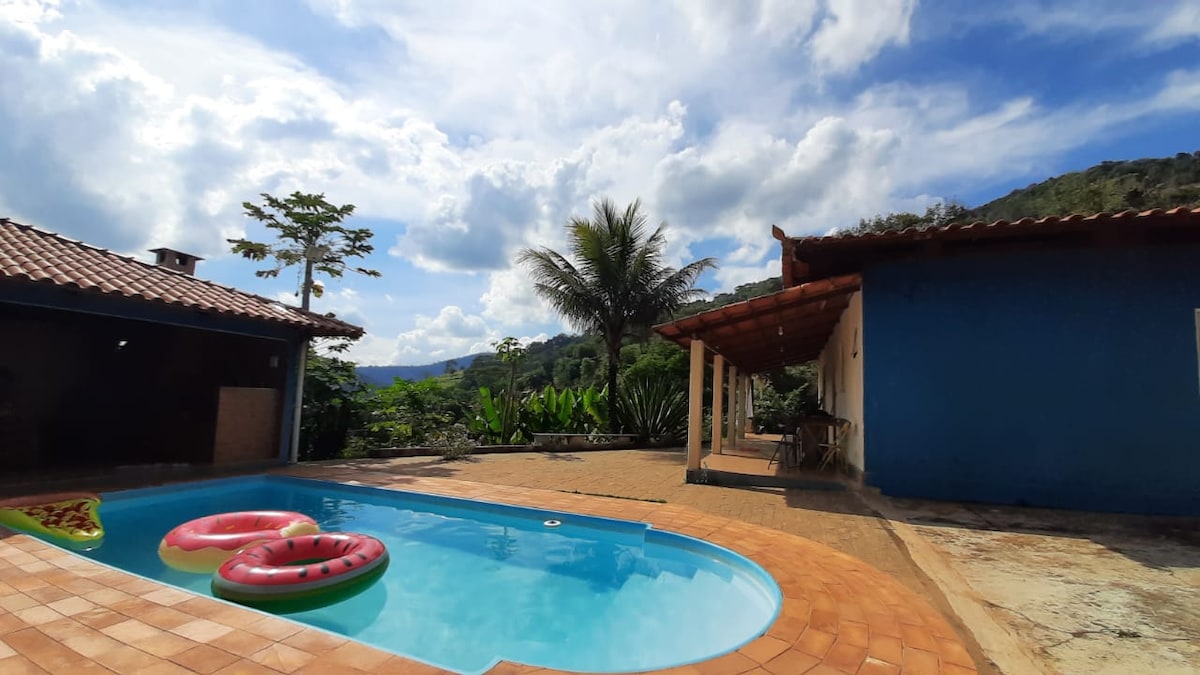 A tranquil outdoor area features a swimming pool surrounded by orange tiles. Two colorful inflatable rings float gently on the water. Lush greenery and palm trees create a natural backdrop, while a blue wall and tiled roof are visible on one side, adding to the peaceful ambiance.