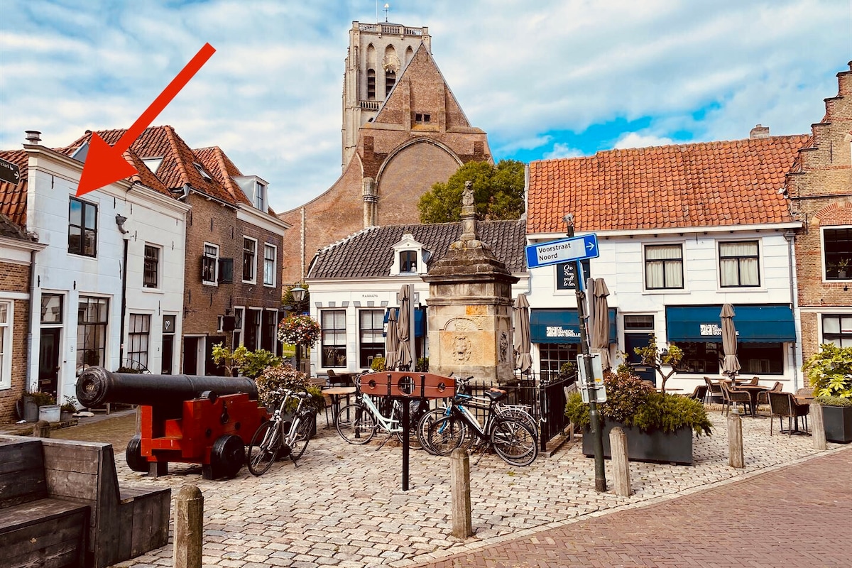 The image features a historic square in Brielle, showcasing cobblestone pathways, charming buildings, and outdoor seating. A prominent cannon and bicycles are visible in the foreground, adding to the sense of the town's heritage. The church tower rises in the background, completing the scene.