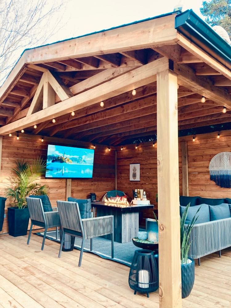 An outdoor gazebo features wood framing with a comfortable seating area and a coffee table. A wall-mounted smart TV is visible, along with decorative plants and ambient lighting from Edison bulbs. The space is designed for relaxation, ensuring an inviting atmosphere for guests.