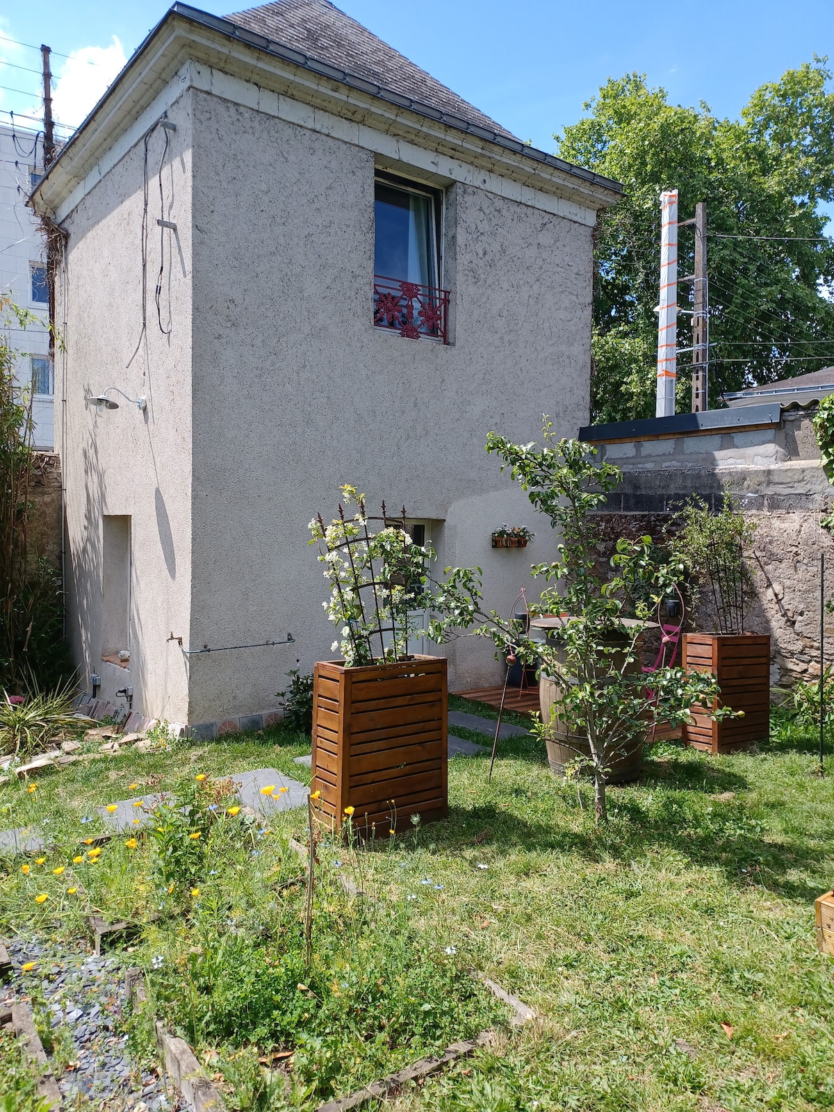 A quaint exterior of a charming old pigeon loft is set against a blue sky. The building features a single window adorned with a decorative railing. Surrounding greenery includes planters with flowers and lush grass, creating a peaceful garden space.