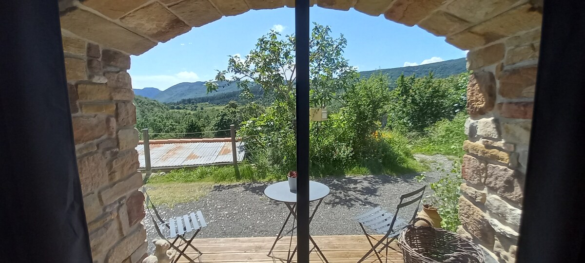 A scenic view is framed by a stone archway, showcasing rolling hills and greenery. A small round table and two chairs sit on the wooden deck outside, inviting relaxation under the clear blue sky.