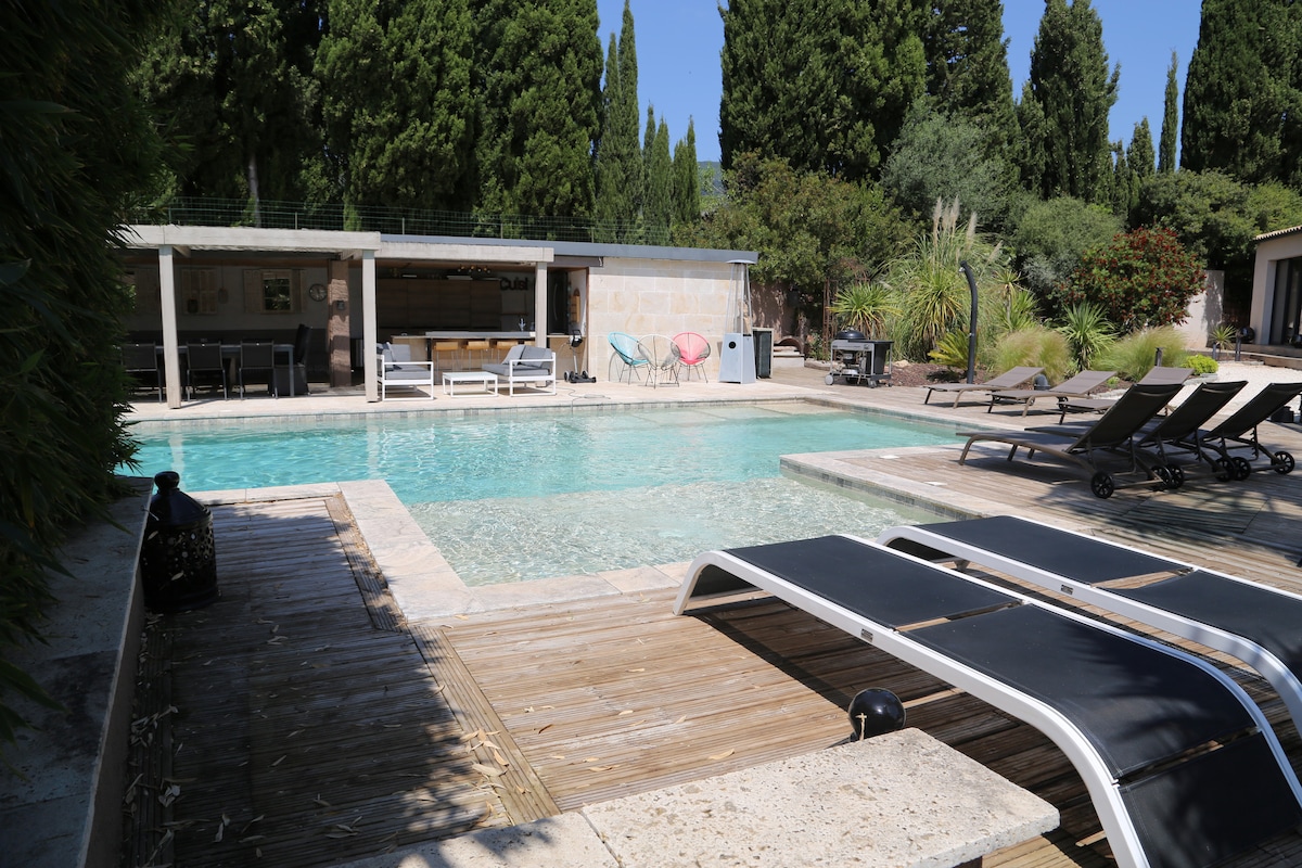 A heated pool is surrounded by a spacious wooden deck and lounge chairs, offering a relaxing outdoor space. An adjacent pool house contains seating and amenities, while lush greenery and tall trees provide privacy and a natural setting.