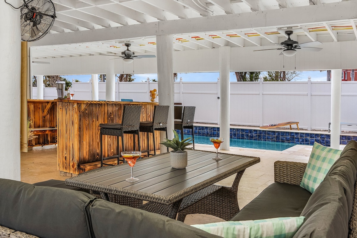 An outdoor seating area features comfortable, shaded lounge furniture surrounding a low table. A wooden bar with high stools is adjacent, while a heated pool can be seen in the background, surrounded by privacy fencing. Ceiling fans provide a cooling breeze.