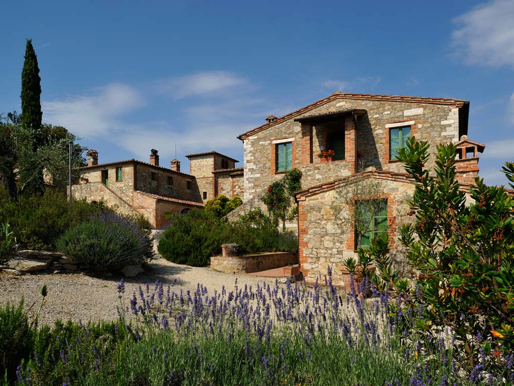 A rustic stone villa sits surrounded by lavender and greenery, showcasing traditional architecture with clay roof tiles and emerald green windows. The pathway leads to additional quaint buildings, under a clear blue sky, inviting a sense of serenity and timeless charm.