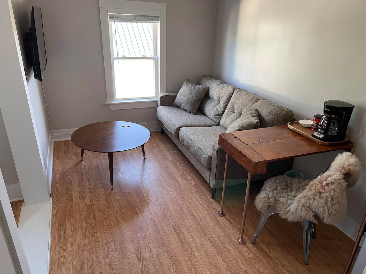 A light-filled living area contains a gray sofa positioned near a window. A round coffee table sits in the center on warm-toned flooring. A wooden desk with a coffee maker and a decorative chair are also present, creating a functional workspace.