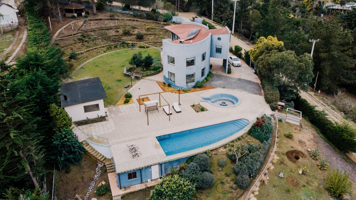 An aerial view of the architect-designed villa reveals a modern structure surrounded by lush greenery. The landscaped garden features a large pool and a terrace area, with outdoor seating and a raised deck. Nearby, additional buildings and pathways are visible in the natural setting.