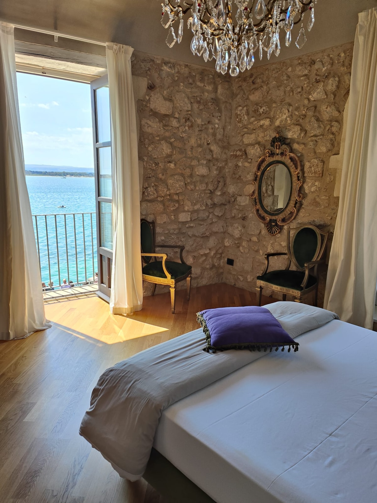 A bright room features a spacious bed adorned with a decorative purple pillow. Natural light floods in from an open window, revealing views of the water. The stone walls add character, complemented by a vintage mirror and two elegant chairs positioned nearby.