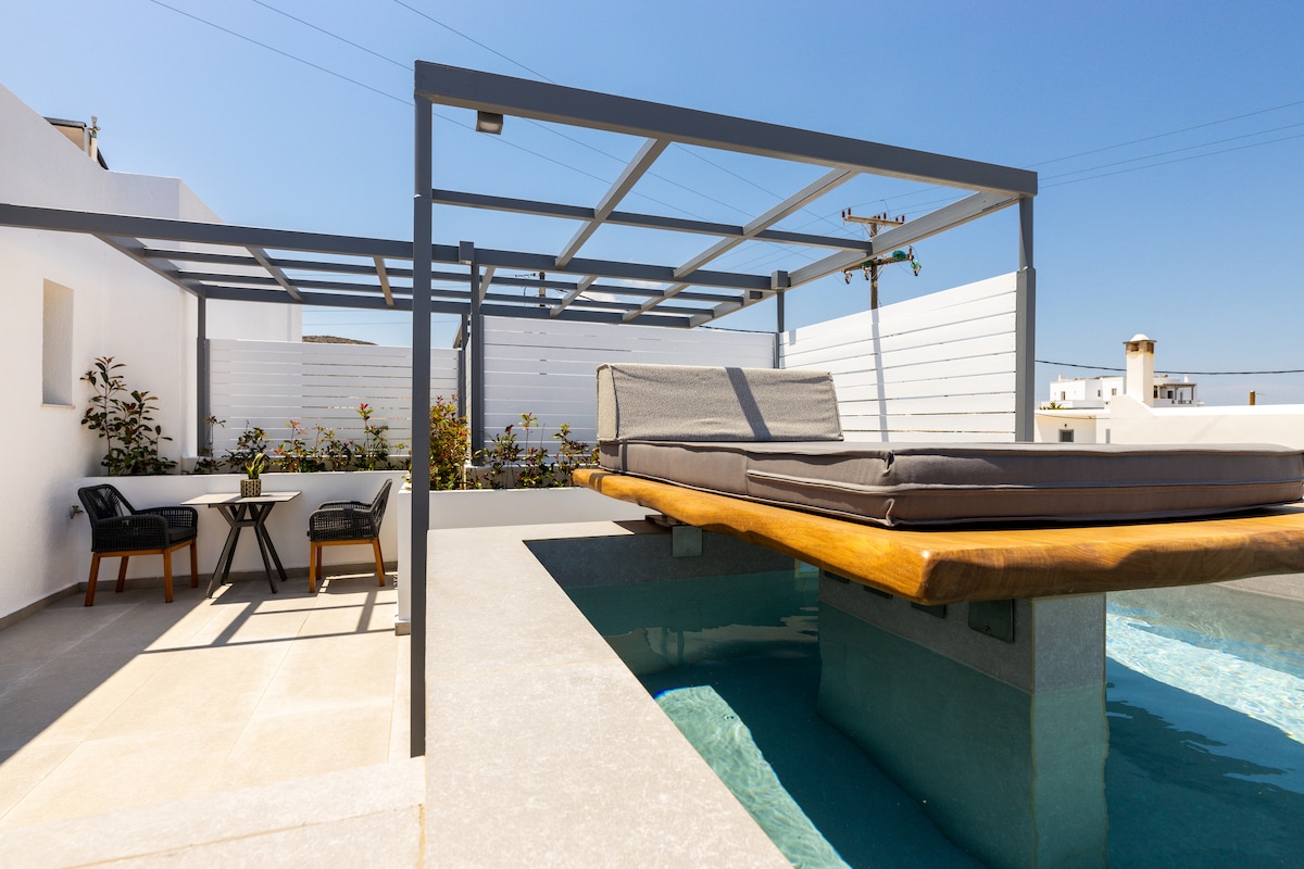 An outdoor area showcases a private jacuzzi surrounded by a stone deck and a sunbathing platform. Two black chairs with a small table sit nearby, sheltered under a modern pergola that provides shade, enhancing the inviting ambiance.