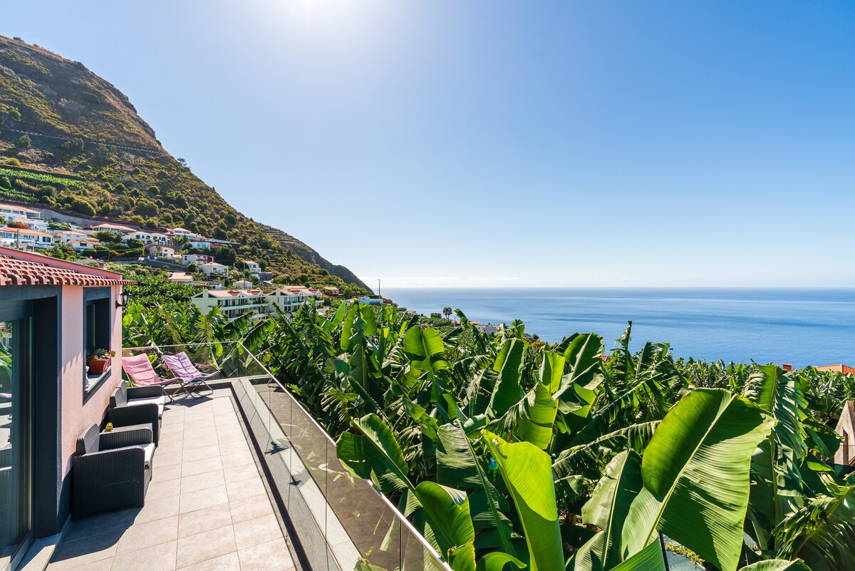 A balcony area overlooks lush greenery and the ocean, creating a serene setting. Comfortable seating is arranged on the balcony, with vibrant banana leaves visible, framed by the distant coastline and mountains that rise against a clear blue sky.
